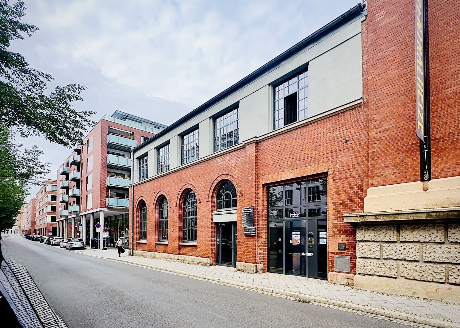 Das Haus des Studios von der Straße aus: Rundbogenfenster, eine Eingangstür und ein verglastes Tor im Erdgeschoss aus Backstein, darüber ein grau verputztes Obergeschoss mit Sprossenfenstern, rechts schließt ein höherer Backsteinbau mit dem Schriftzug „Zentralheize“ an